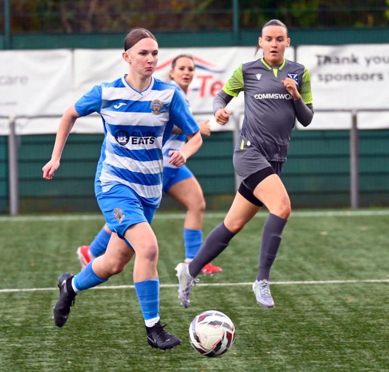 Three women playing football. A womanin a blue striped top is dribbling the ball with a player in a different strip nearby