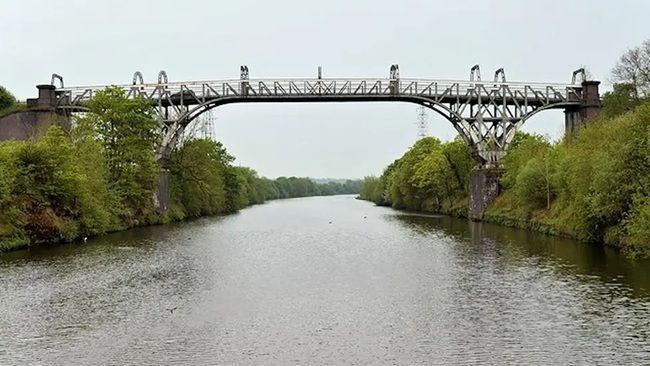 A metal bridge straddles two wooded banks over a large river on a clear day.
