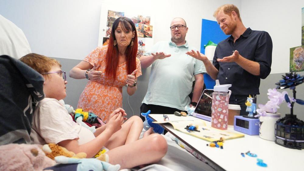 Prince Harry meets a young child in hospital. He stands by the bed with the boys parents.