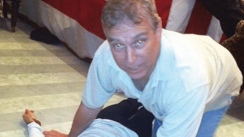 A grey-haired man dressed in a white polo shirt and blue jeans looks directly at the camera. He is on all fours directly over an unidentified female who is laying on the floor.