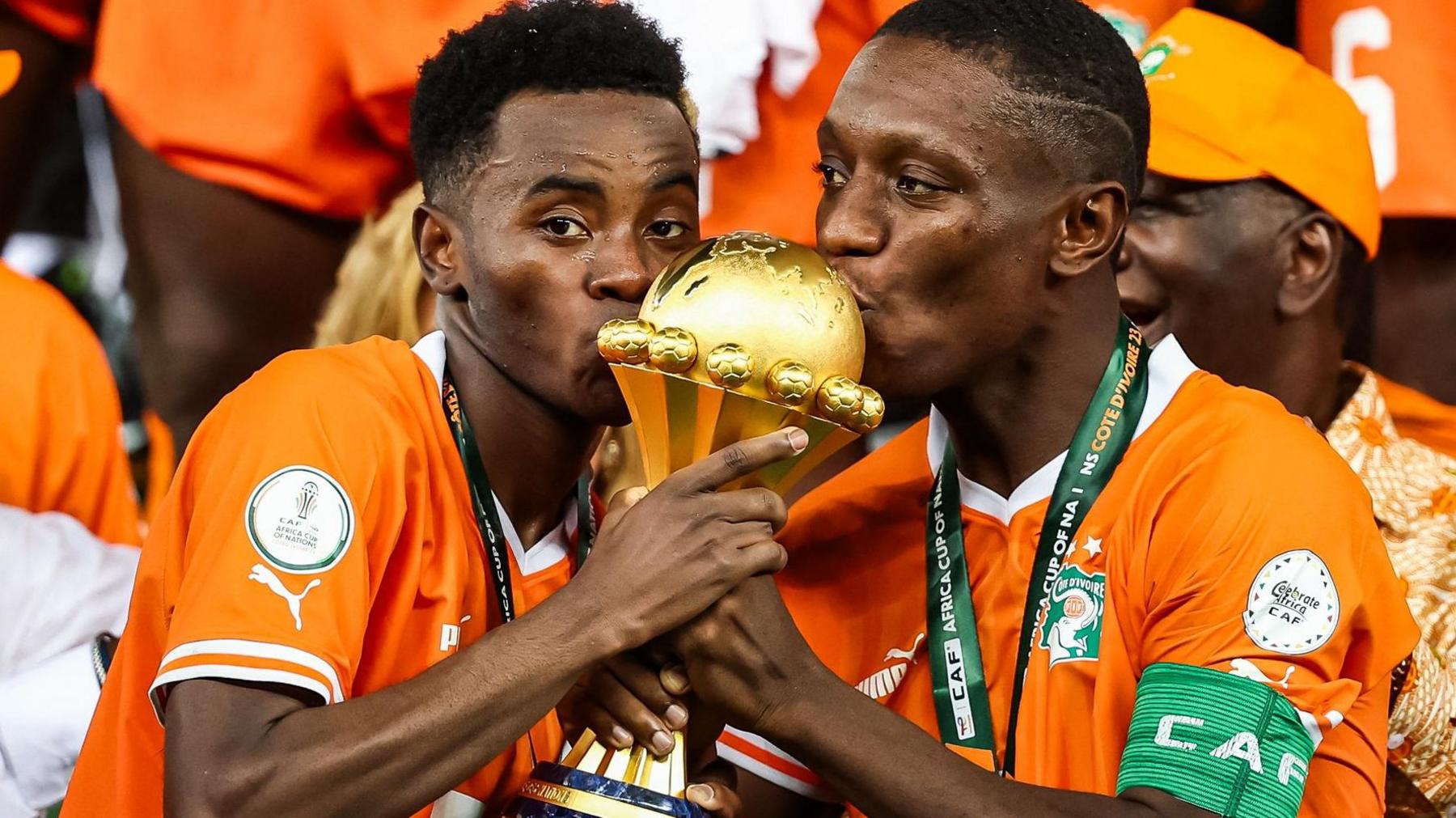 Simon Adingra and Max-Alain Gradel seen from chest up, both wearing bright orange Ivory Coast football shirts with medals around their necks, hold the golden Africa Cup of Nations trophy between them and kiss the top of it. Adingra is looking directly into the camera while Gradel casts his eyes to the left of the screen. The top of the trophy which they are kissing is the upper hemisphere of a globe which is submerged into a long, curving handle