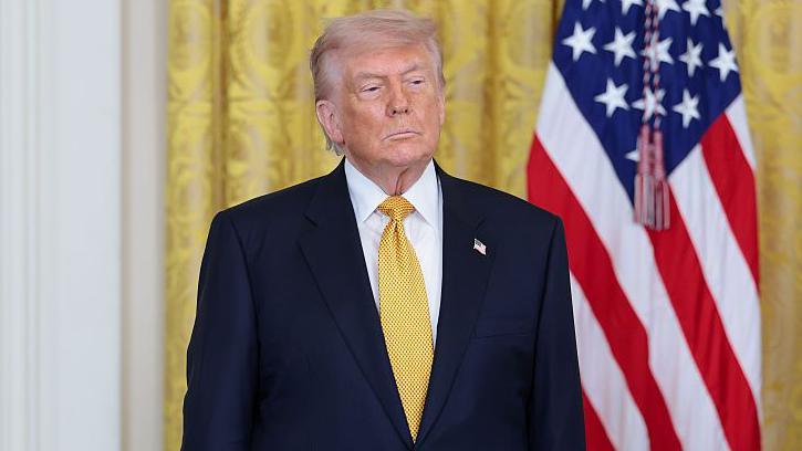 Donald Trump, in a dark navy suit and yellow tie, stands with his hands at his sides and a neutral expression. Behind him are gold curtains in the White House and an American flag.