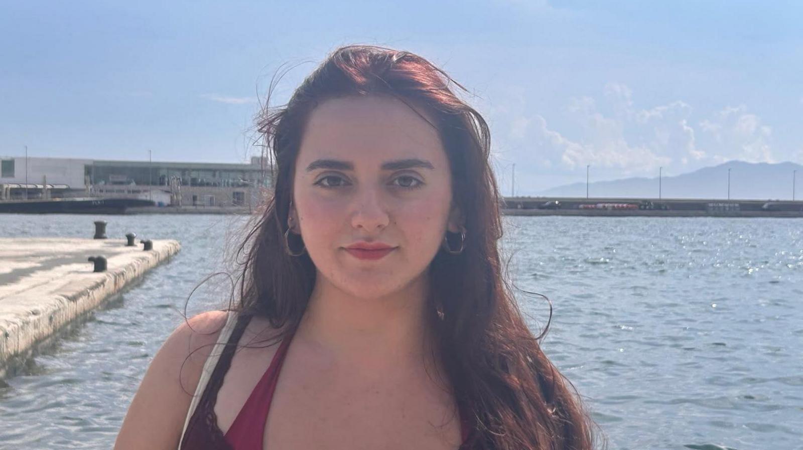 The image is of a young woman with long, dark brown hair falling over her left shoulder. She is looking directly into the camera with a calm expression. She has dark eyebrows, dark eyes, and is wearing a brown top over a burgandy swimsuit, and has small, metallic hoop earrings. Behind her is a wide expanse of calm water with a concrete pier jutting out and several bollards along the edge. In the distance is a long, flat-roofed building and beyond that is a low mountain.