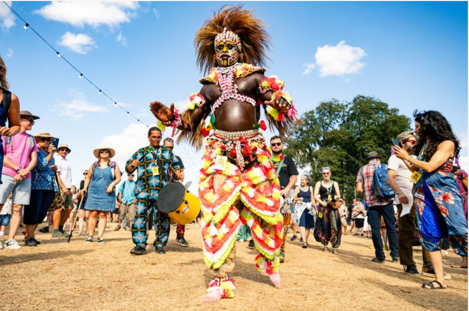 A man in a colourful outfit dances as a man plays a drum behind him