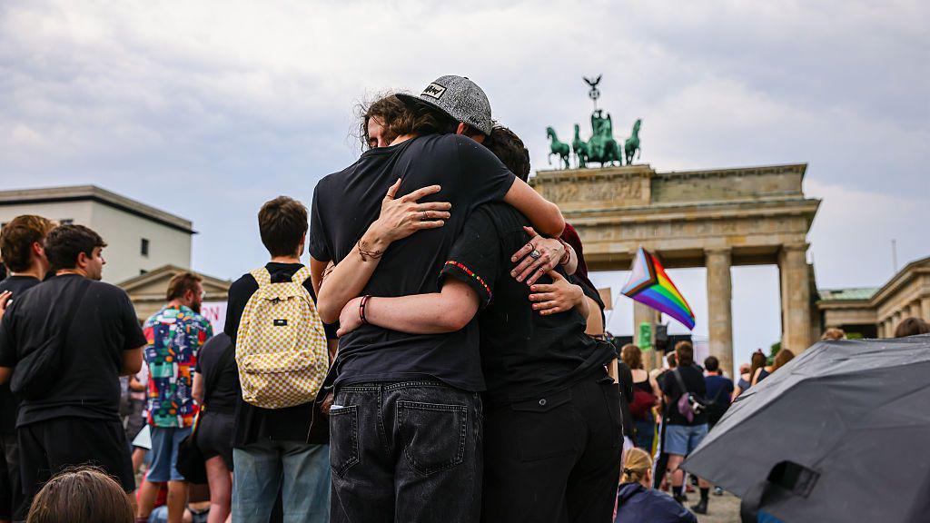 Three people hug each other with a rainbow LGBT flag visible in the background and the Brandenburg Gate behind