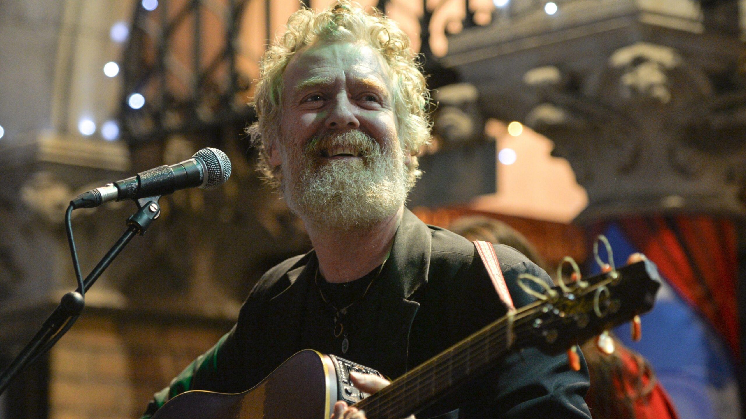 Glen Hansard smiling as he plays guitar outside a Dublin theatre on Christmas Eve night 2019.  He is a man with greying ginger hair and a matching beard.  He is wearing a dark green blazer over a dark t-shirt