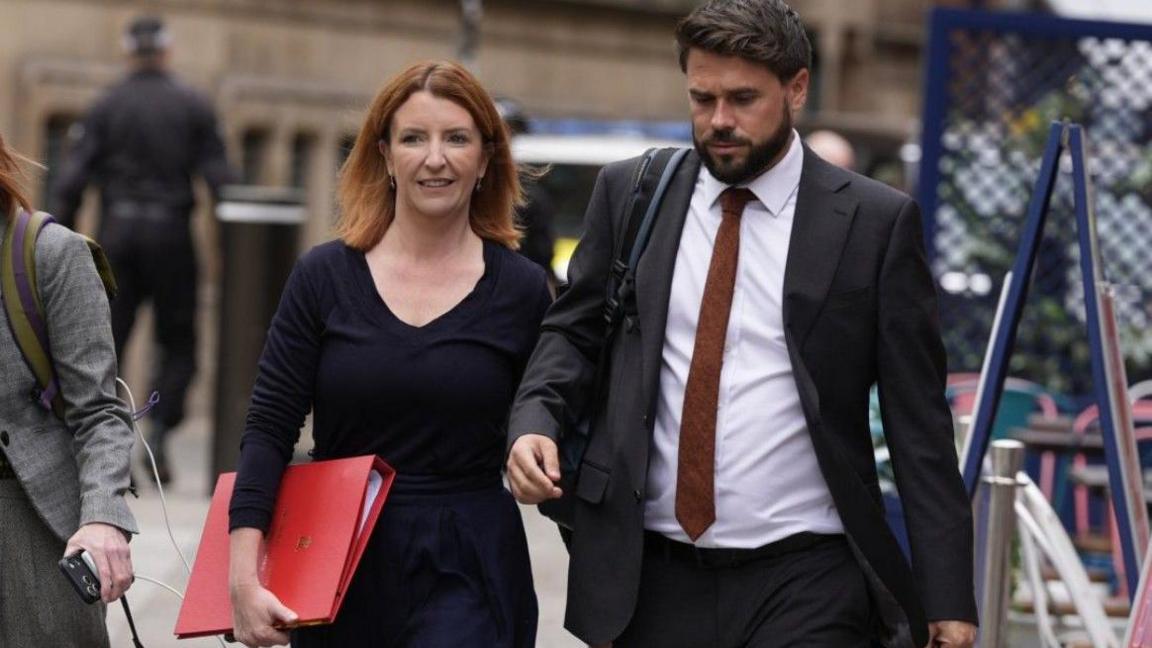 Louise Haigh is walking down the street next to an aide. She is carrying a  large, scarlet government folder and wears a smart black top and black trousers. Her hair is ginger and shoulder length, and she wears it loose, along with a pair of small earrings.