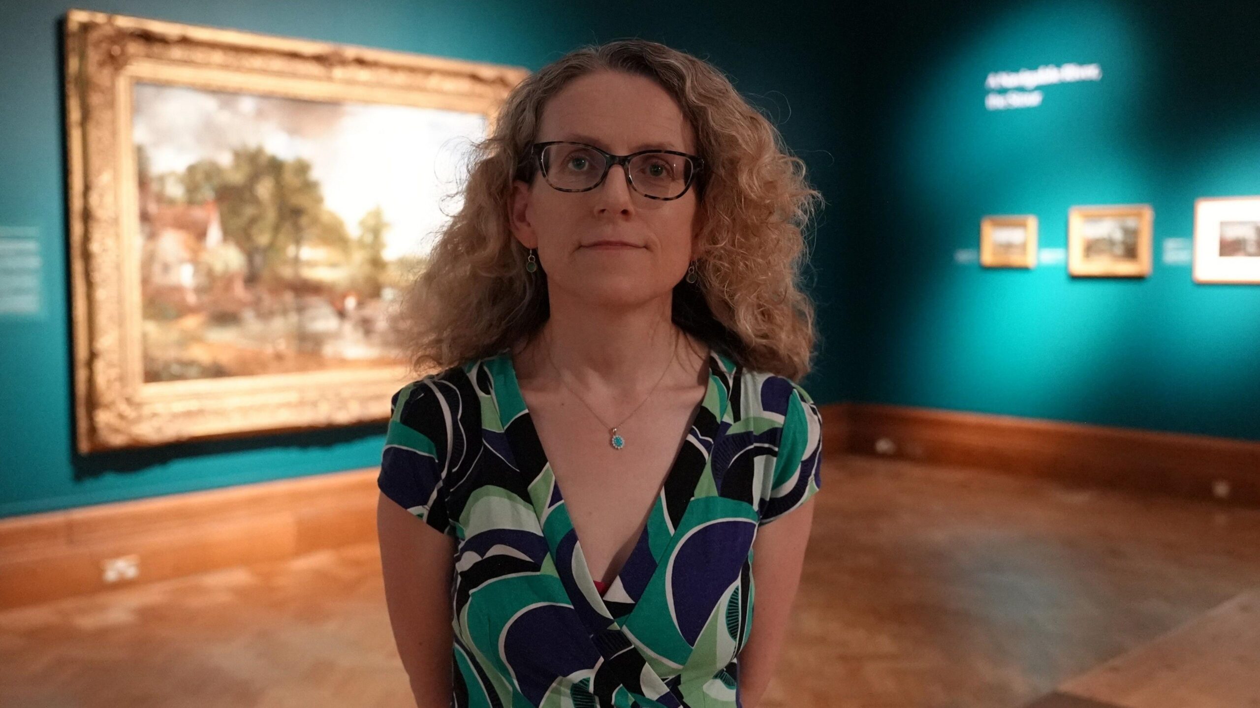 A woman wearing glasses stands in the middle of an art exhibition to pose and smile at the camera. She has long curly blonde hair and is wearing a green and blue dress.