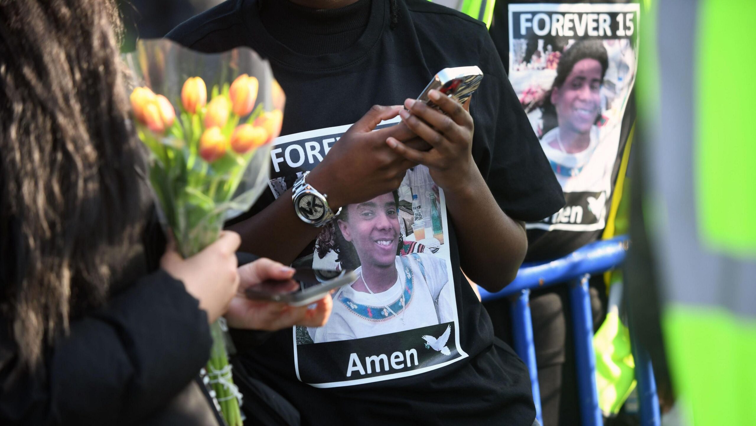 Close-up  of people wearing tshirts with Forever 15 Amen printed on them. One person is holding a bunch of flowers and another is holding a mobile phone.