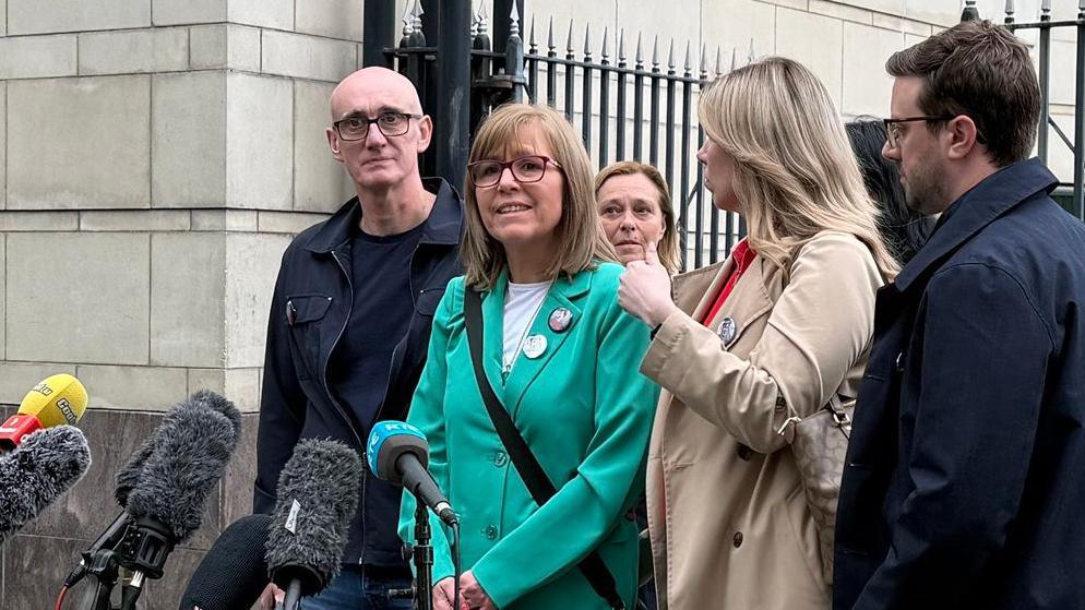 Lyra McKee's sister Nichola is wearing a green jacket and is speaking into a microphone outside court. She has sandy coloured hair and glasses. Other family members stand beside her