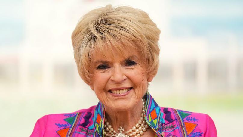 Gloria has short blonde hair and is wearing a bright patterned jacket with a pearl necklace.
