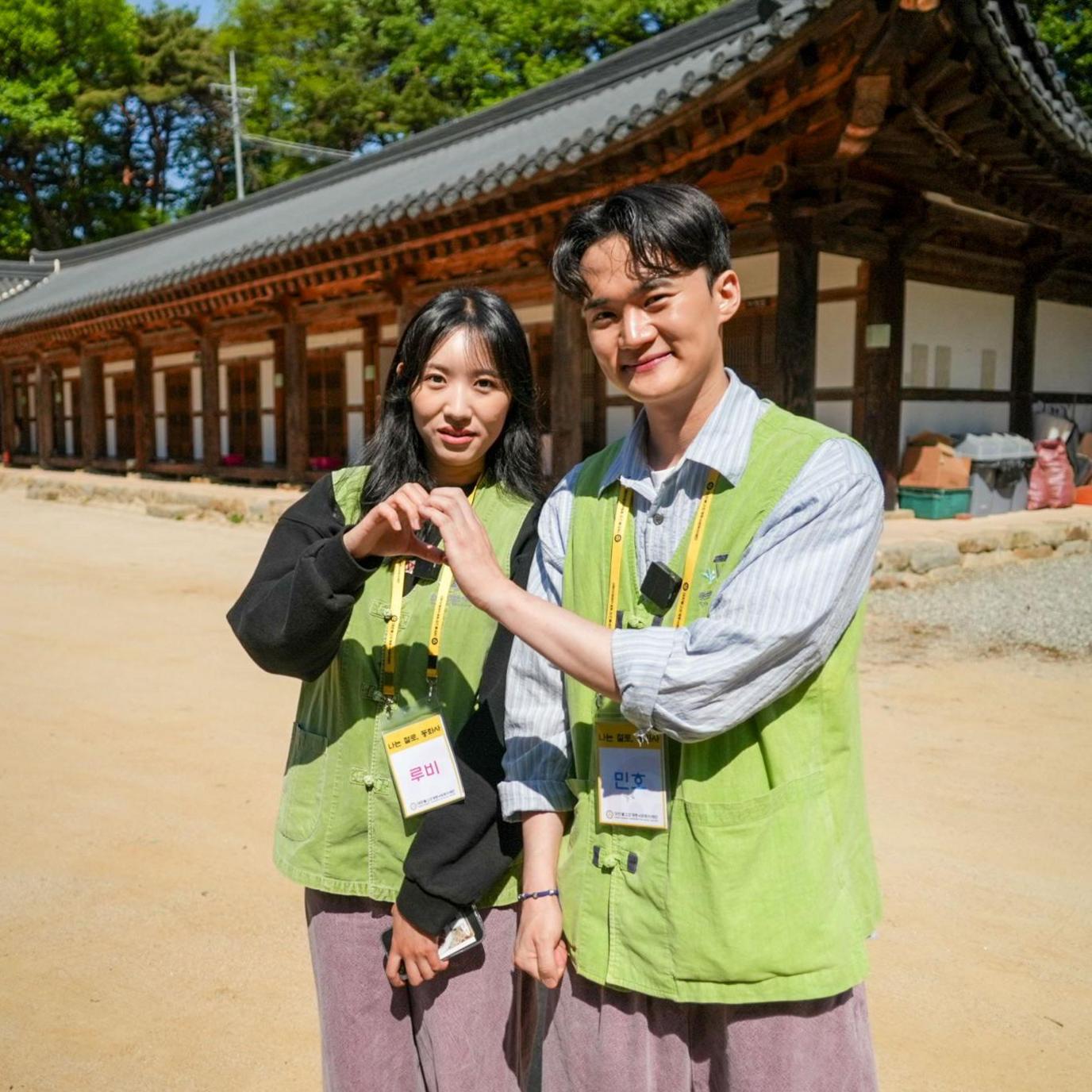 Two participants - Ruby and Minho - wearing light green vests and event lanyards stand in a courtyard, joining hands to form a heart shape.