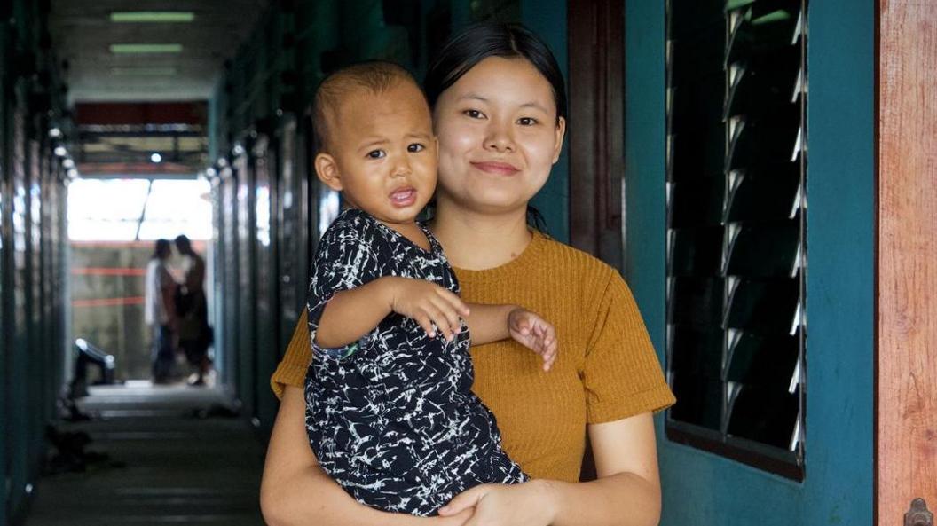 Young Burmese woman wearing a brown shirt, her hair tied, holding a baby wearing a black and white shirt and shorts, with an open door and a corridor in the background in Mae Sot, Thailand, that borders Myanmar