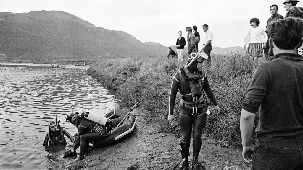 Rescue divers at the edge of the Mawddach estuary in July 1966, watched by onlookers, during the search for survivors after the Prince of Wales pleasure boat sank. The divers are in full wetsuits and have scuba diving equipement, air tanks, masks and snorkels.