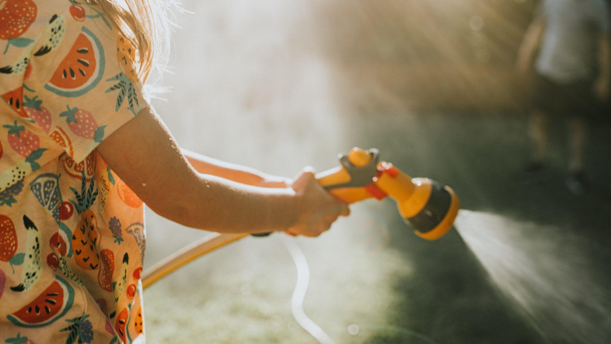 A girl wearing a patterned top holds a yellow garden hose with an attachment. She sprays water from the hose attachment. A man stands in the top right background of the picture.