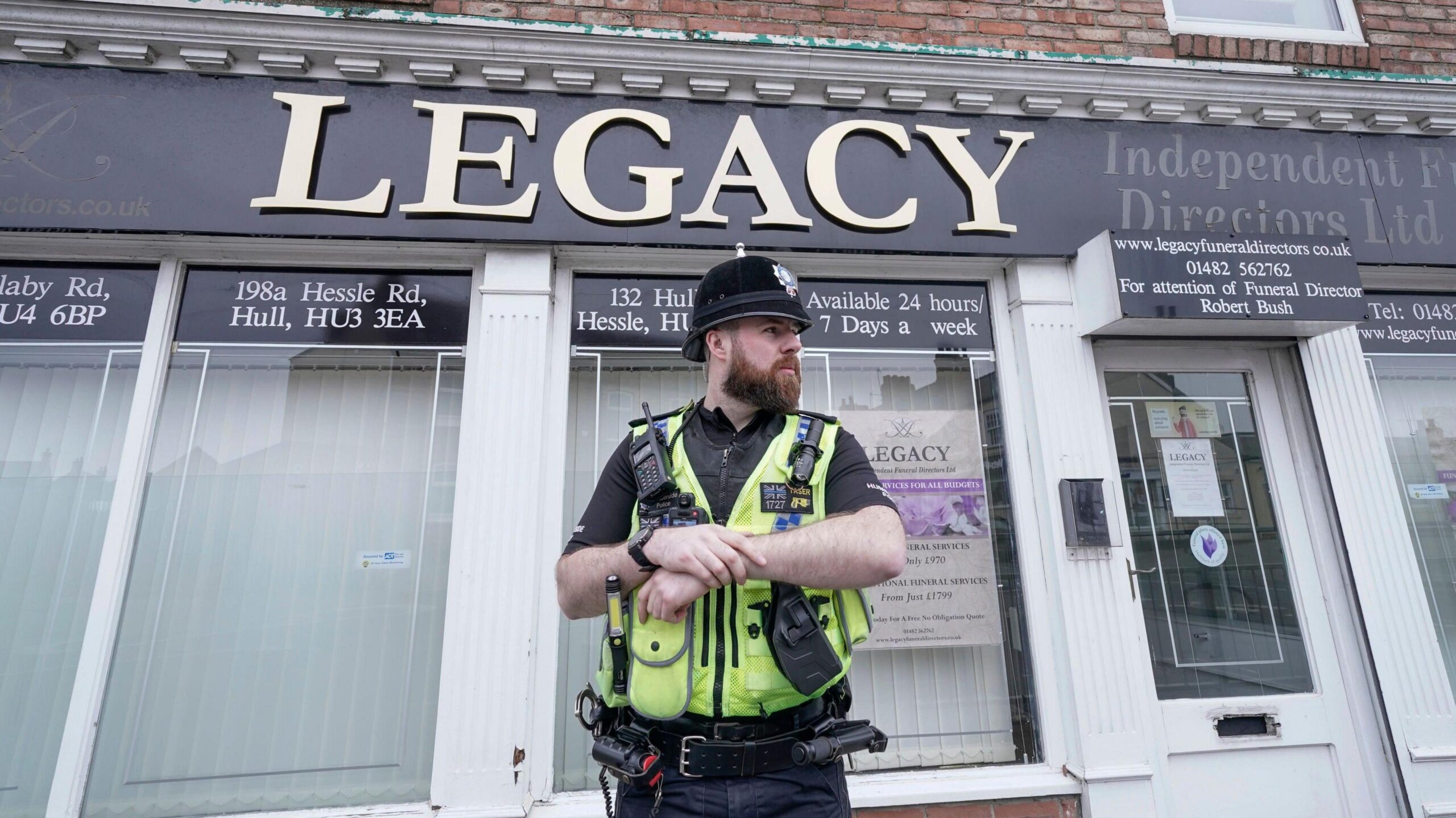 A male police officers stands outside a funeral parlour. He is wearing a black helmet and a yellow, high-vis vest over a black uniform. The funeral parlour has large glass windows and closed white blinds. A sign has gold letters on a black background, reading "Legacy". Smaller signs advertise the various branches of the business, with addresses around Hull.
