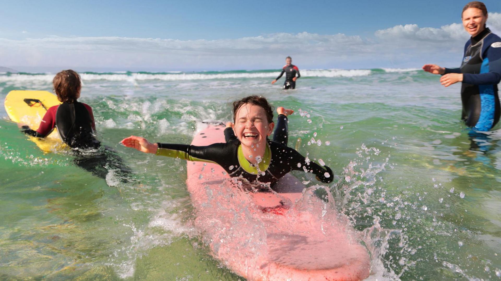 A family of four wearing wetsuits and learning to surf. One young child is lying on a red surfboard, facing the camera and grinning; the other child is holding on to a yellow surboard with their back to the camera. The parents are standing in the water, in the distance.