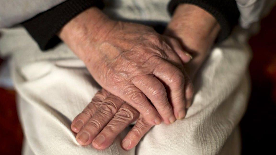 A close-up shot of the hands of an elderly woman. She has them folded on her lap, neatly trimmed and without nail varnish. She is wearing cream linen trousers.