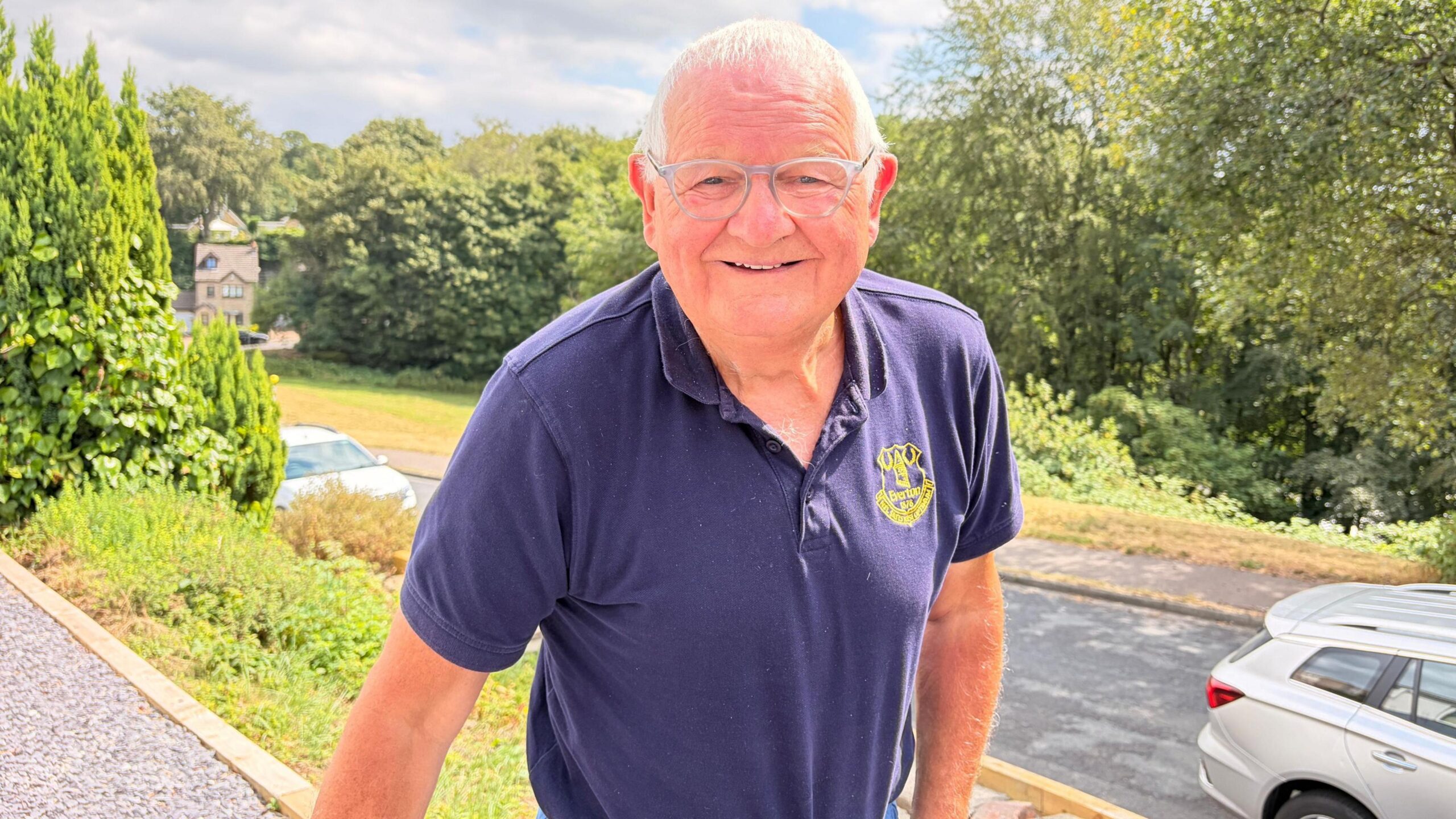 A man in a navy blue T-shirt and wearing glasses stands in front of a quiet tree-lined street, smiling and looking relaxed.
