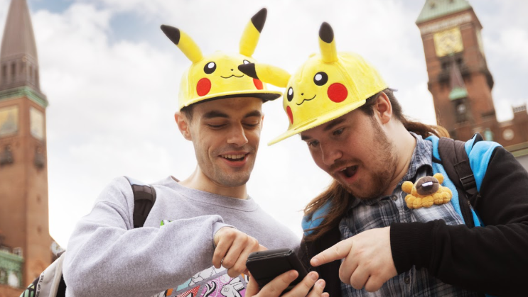 Two Pokémon Go fans wearing hats shaped like the yellow Pokémon character Pikachu are pointing and looking at a phone, in shock.