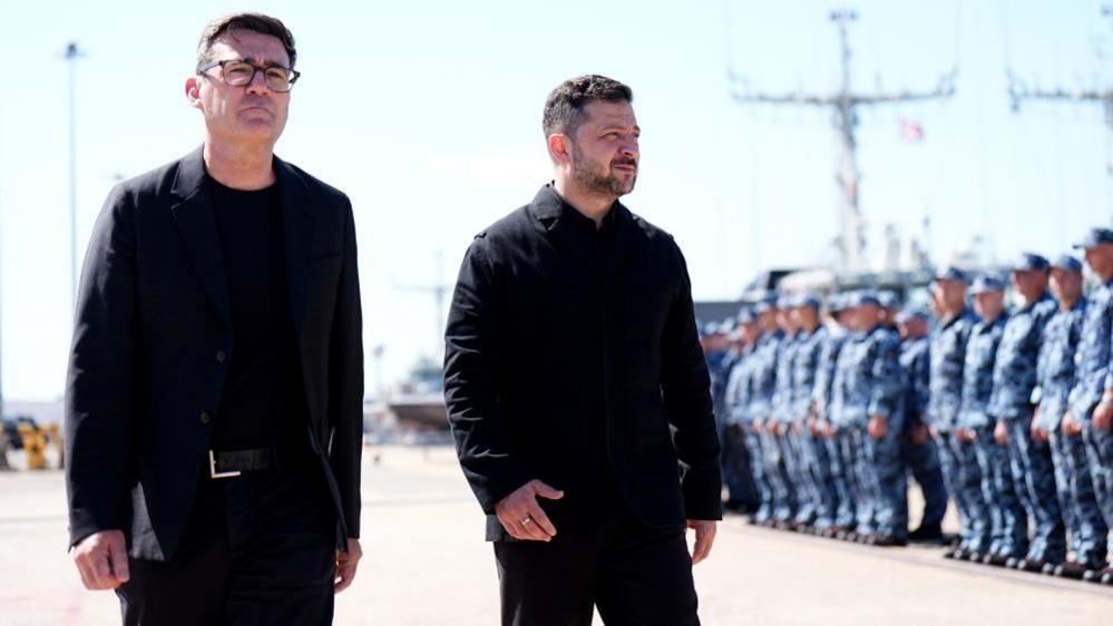 Andy Burnham and Volodymyr Zelensky walk next to each other at a naval base. Both men are dressed in black and have short, dark hair. Burnham is wearing glasses. Military personnel can be seen in the background.