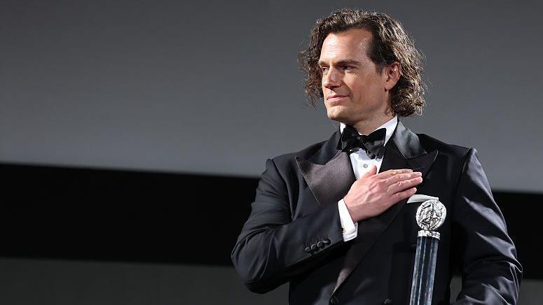 Henry Cavill wears a suit and bowtie as he stands on-stage holding a tall, cylindrical award. He places his free hand flat across his chest and smiles appreciatively at someone off-stage.