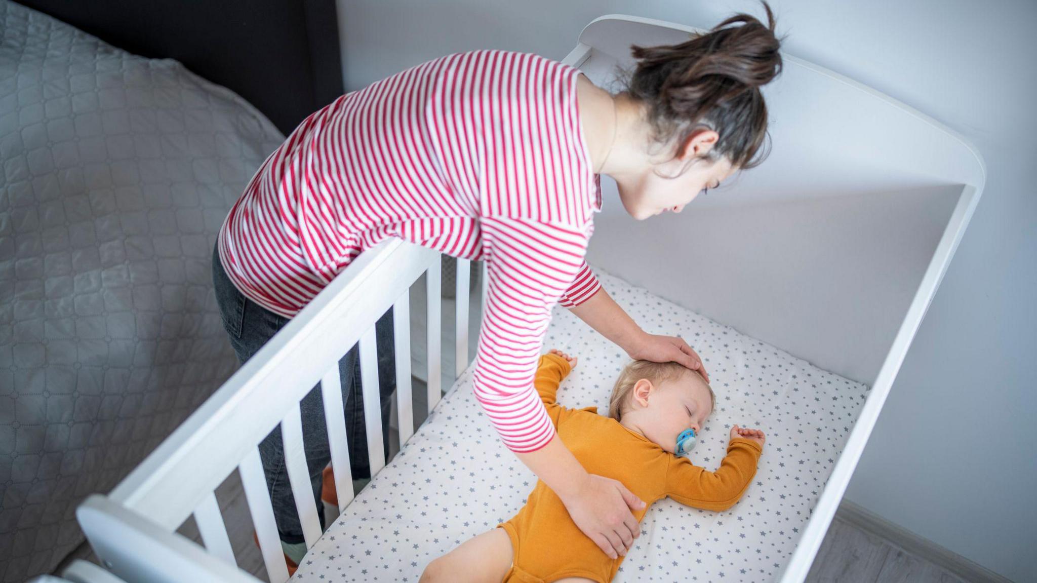 A woman leaning over a white cot, adjusting a baby lying on their back on a light-patterned mattress. The adult is wearing a red-and-white striped long-sleeved top, and the baby is dressed in an orange outfit. The scene is indoors, with a bed visible beside the cot.
