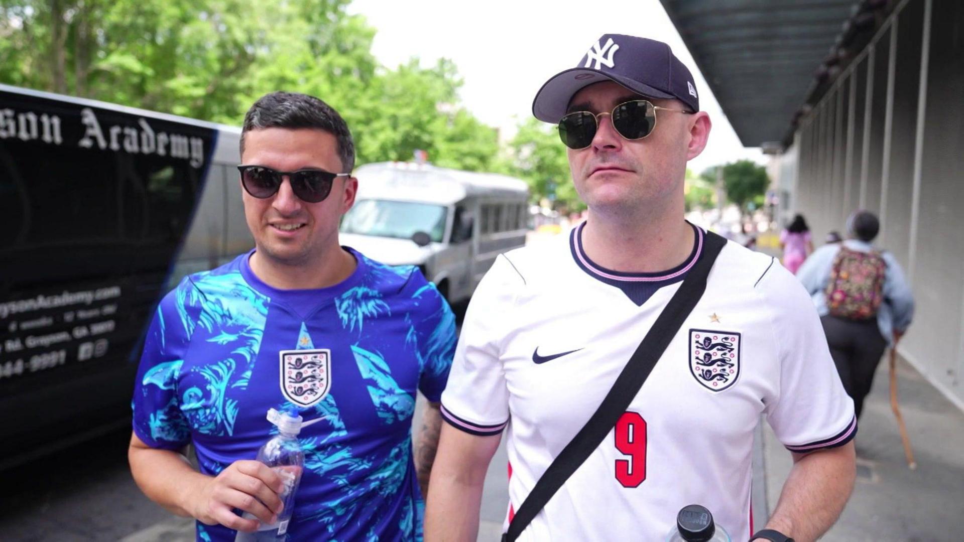 A man in a blue shirt and wearing sunglasses, he is holding a bottle. He is standing next to a man, also wearing sunglasses, in a white shirt with a red number nine printed on his shirt.