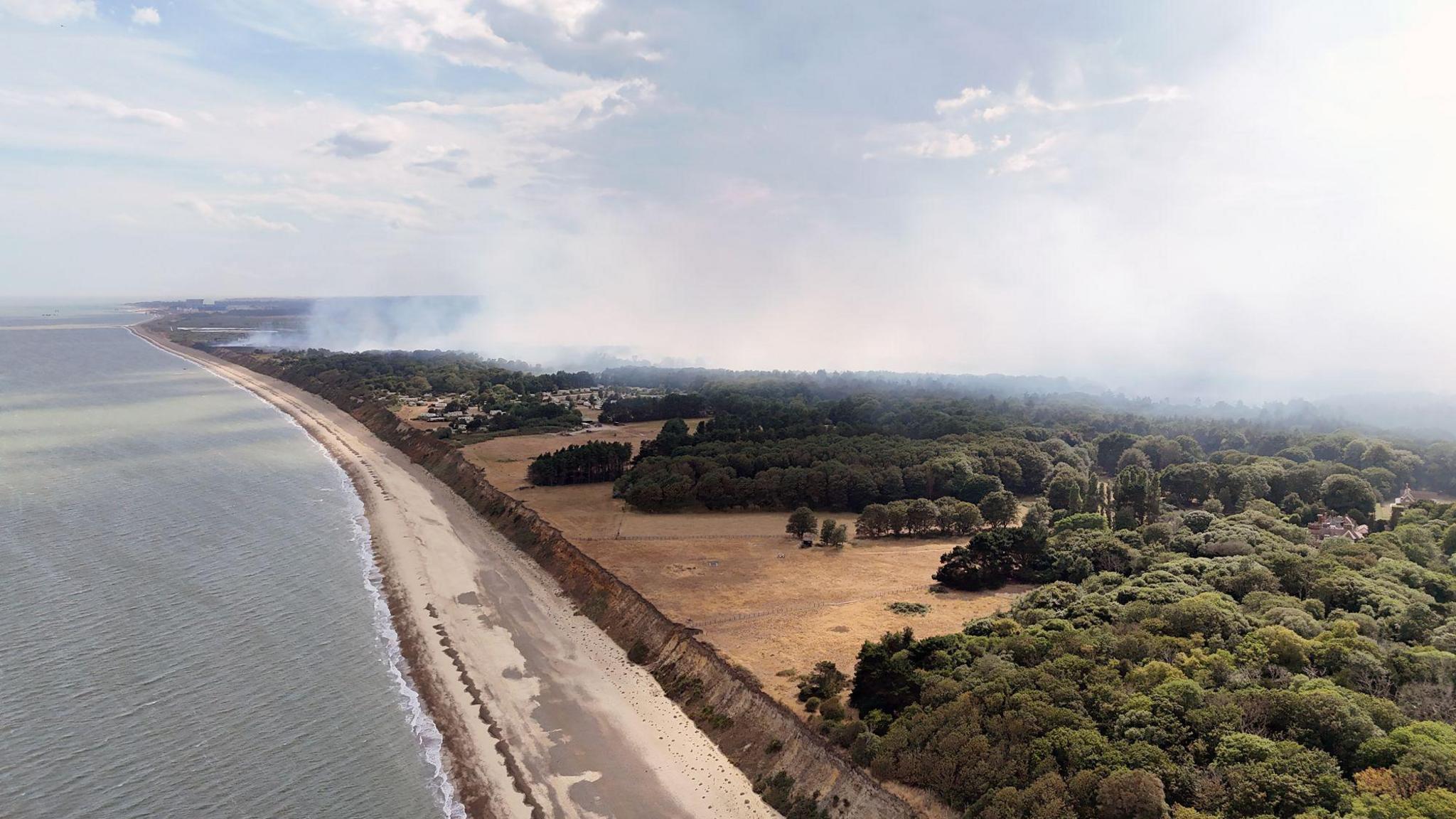 Aerial image shows the sea on the left of the frame with trees and heathland and a large pall of smoke.