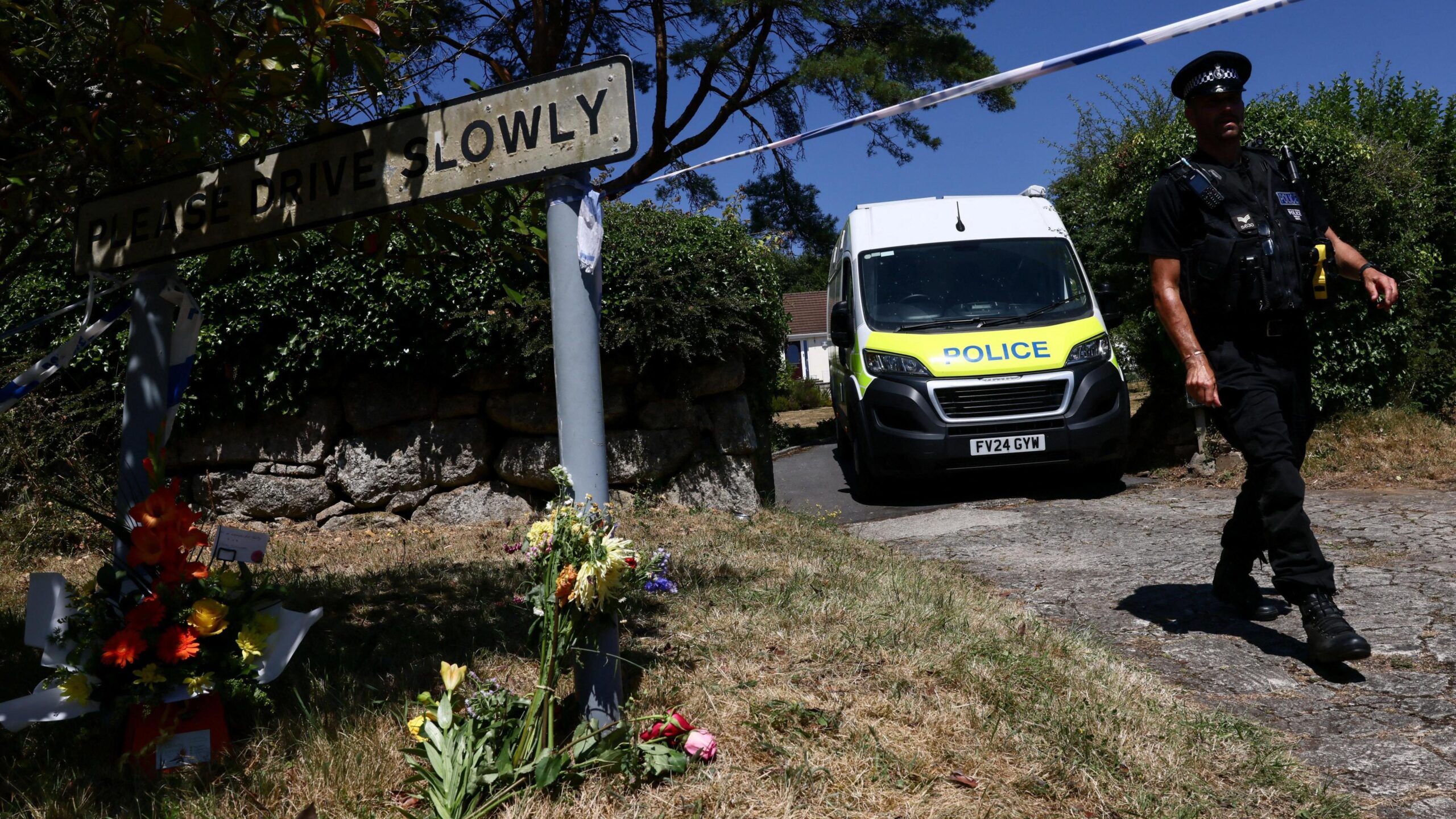 Flowers are pictured on the ground with a police officer walking down a driveway beside