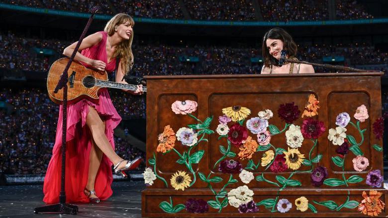 Taylor Swift and Gracie Abrams perform on stage during "Taylor Swift | The Eras Tour" at Wembley Stadium on June 23, 2024 in London. Taylor wears a flowing bright pink dress and holds an acoustic guitar, kicking out her right leg. Gracie sits behind a piano painted with flowers, she has short brown hair and brown eyes.
