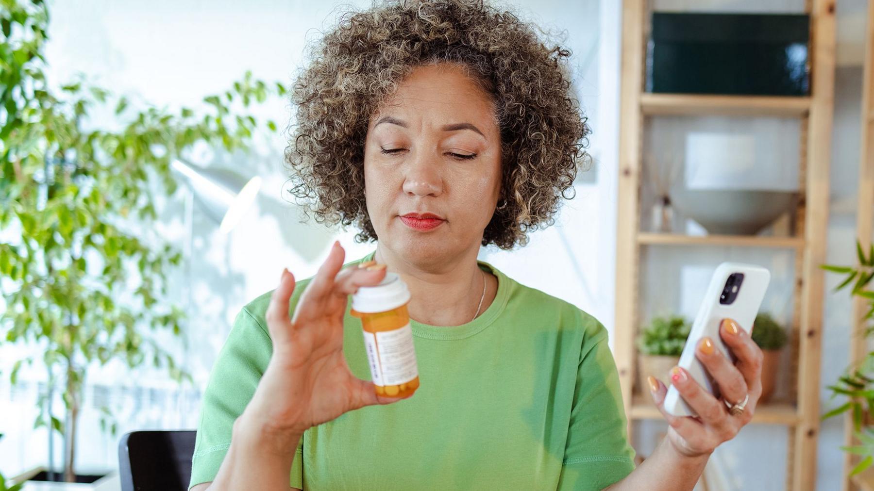 A woman in a bright green T-shirt holds a bottle of pill in one hand and a phone in the other and looks at the bottle of pills