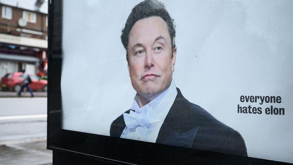 A poster of Musk wearing a suit and bow tie on an advertising display panel in London, with "everyone hates elon" written on it.