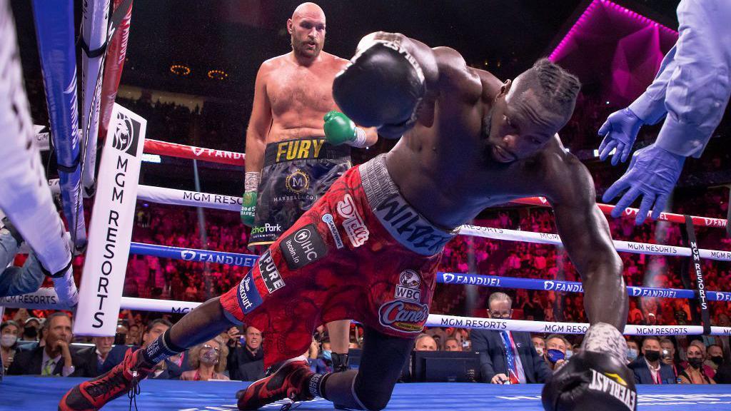 Tyson Fury watches as Deontay Wilder falls to the canvas in a boxing ring