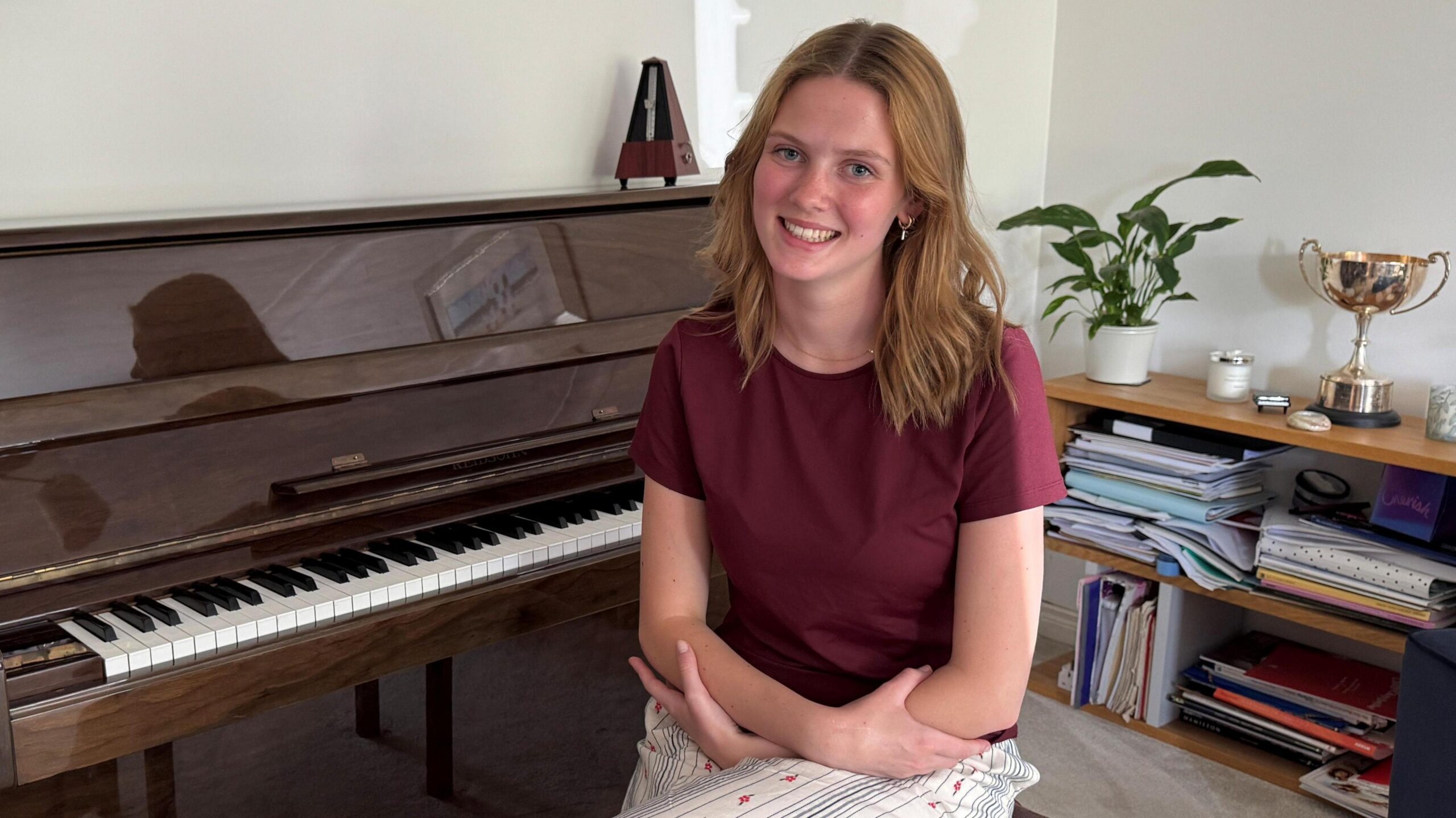 Freya Terris is a young woman with shoulder length light brown hair, which is hanging in loose waves around her face. She is smilling at the camera. She is wearing a burgundy t-shirt and is sitting on a piano stool with her arms crossed. She is sitting in front of a glossy brown piano, and there is a metronome, a plant and a large trophy seen sat around her.