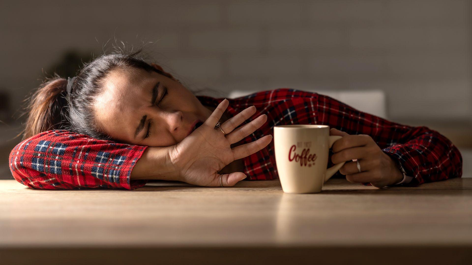 A woman in a red checked shirt rests her head on a table next to a cup displaying the word 'coffee'
