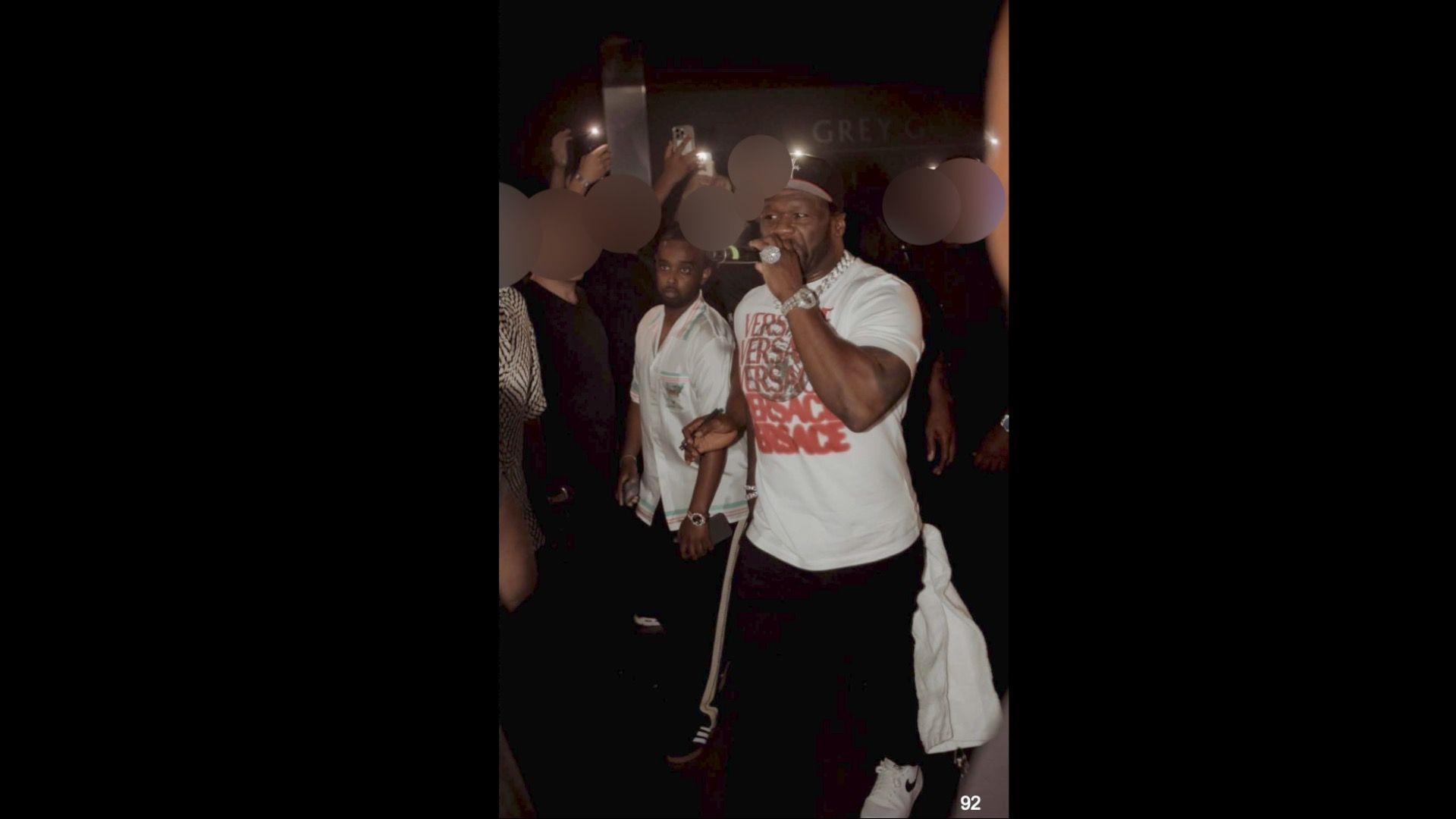 Hassan, wearing a silky button up shirt, stands next to 50 Cent, who is holding a microphone.