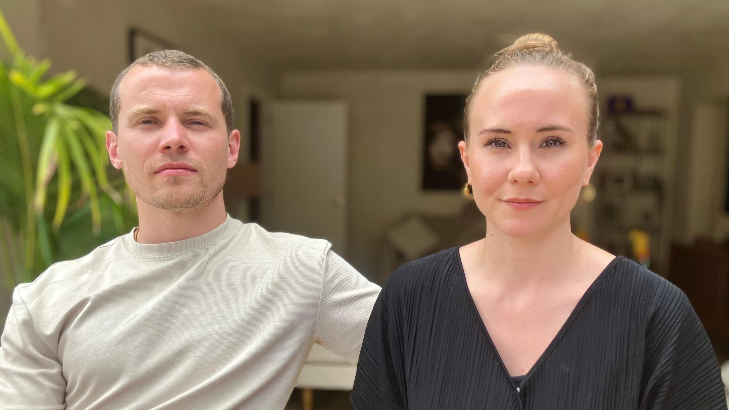 A man and woman sitting together. The man wears a pale T Shirt and the woman a black V neck T Shirt and they are sitting in a living room with plants and pictures on the wall.