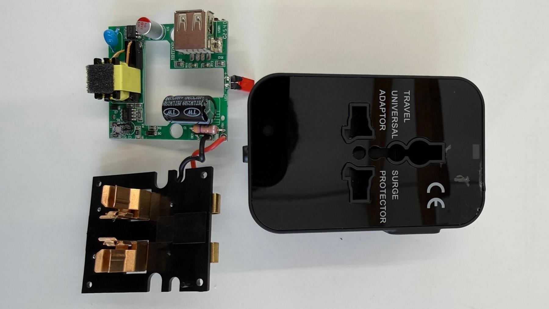 A circuit board and small plastic cover on a table beside a black, plastic travel adaptor. Text on the adaptor case, next to its sockets, reads "TRAVEL UNIVERSAL ADAPTOR" and on the other side: "SURGE PROTECTOR". A "CE" certification standard symbol is also displayed on the casing. Copyright