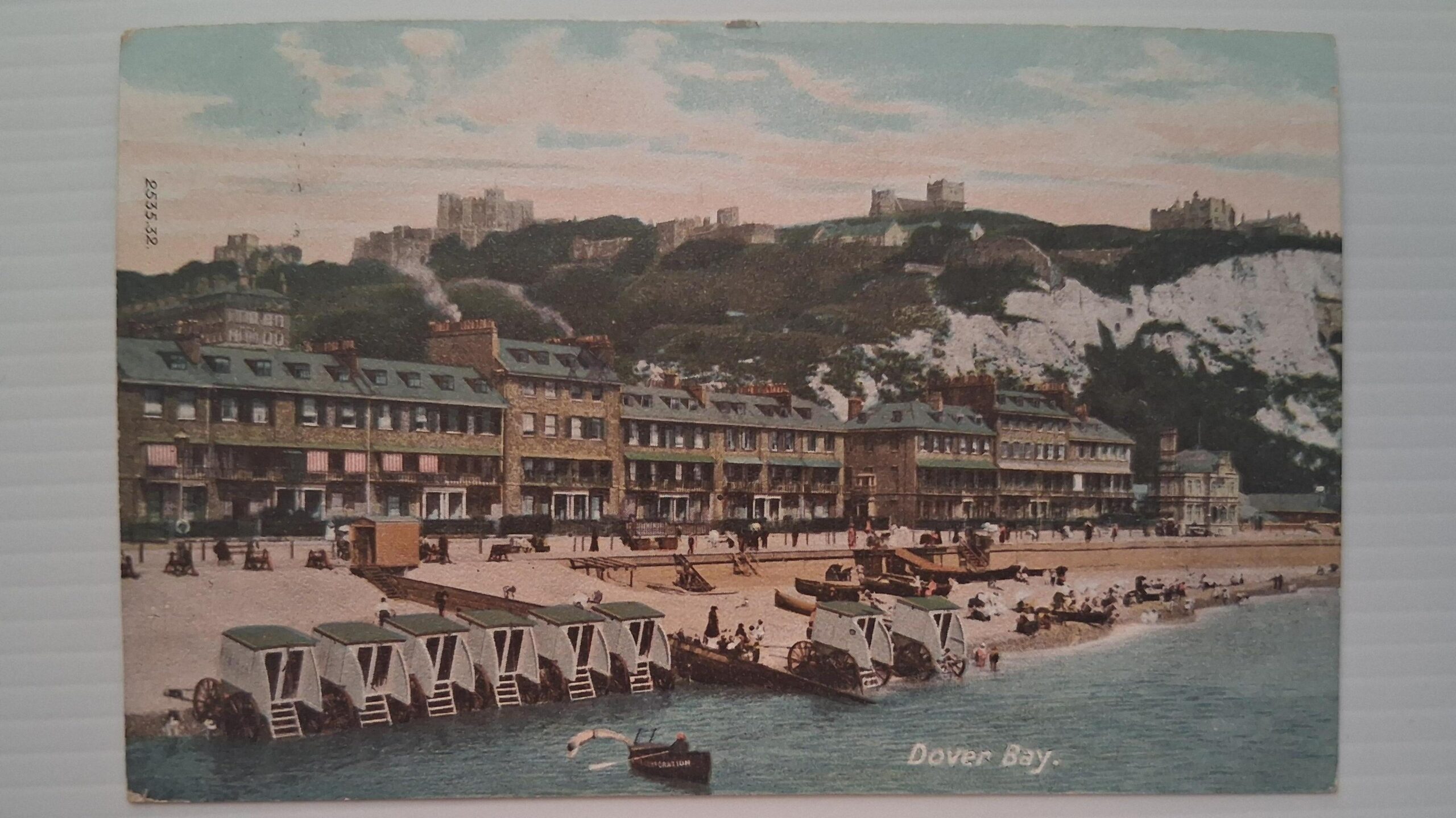 A postcard with an illustration of a beach with buildings, the white cliffs of Dover and Dover Castle. The illustration is labelled Dover Bay.