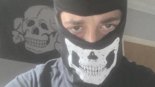 Alfie Coleman wearing a black balaclava with a skull on it. Only his eyes can be seen. Behind him a black flag with a white skull and crossbones is pinned to a wall.