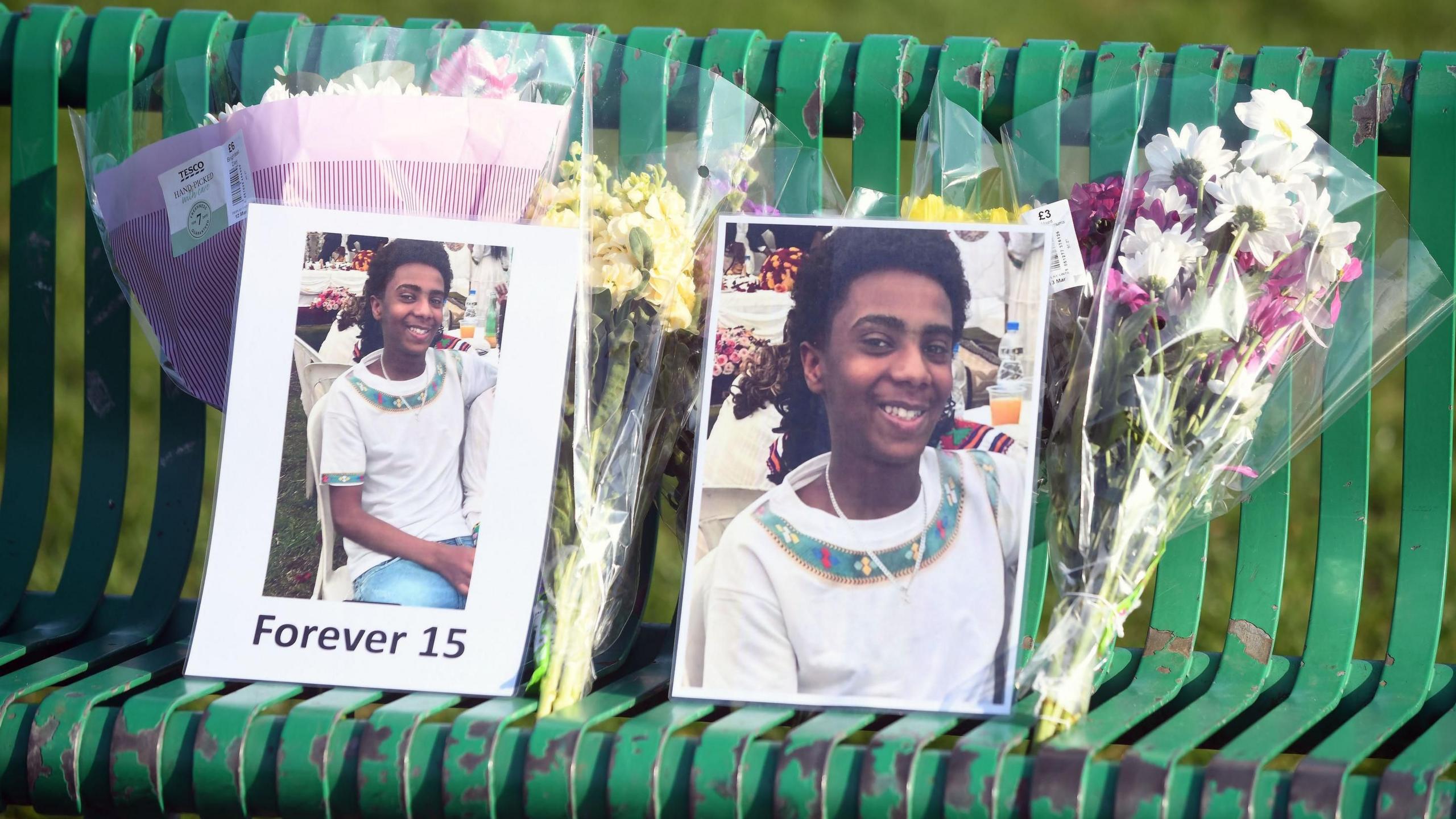 Tributes to Amen on a green bench. There are pictures of him smiling and bunches of flowers.
