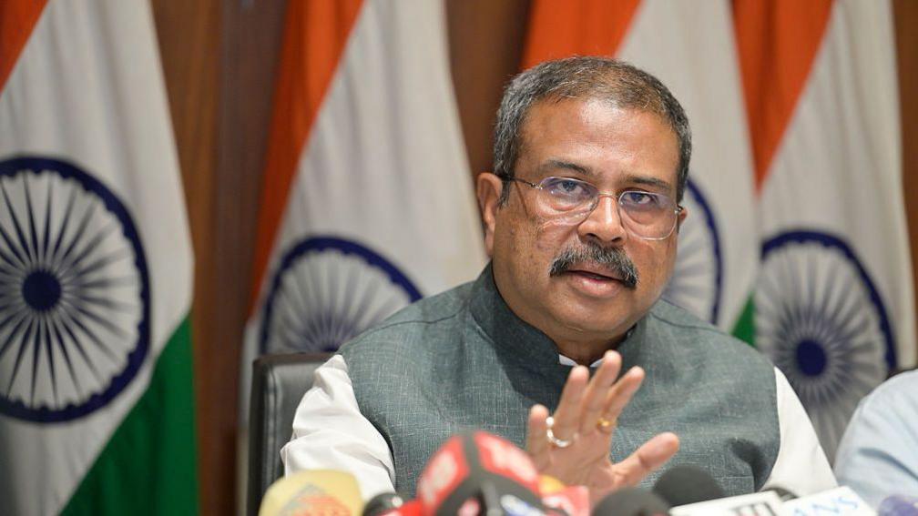 Union Minister of Education Dharmendra Pradhan addresses a press conference at his residence, pictured with one hand up and Indian flags in the background, wearing a grey and white garment