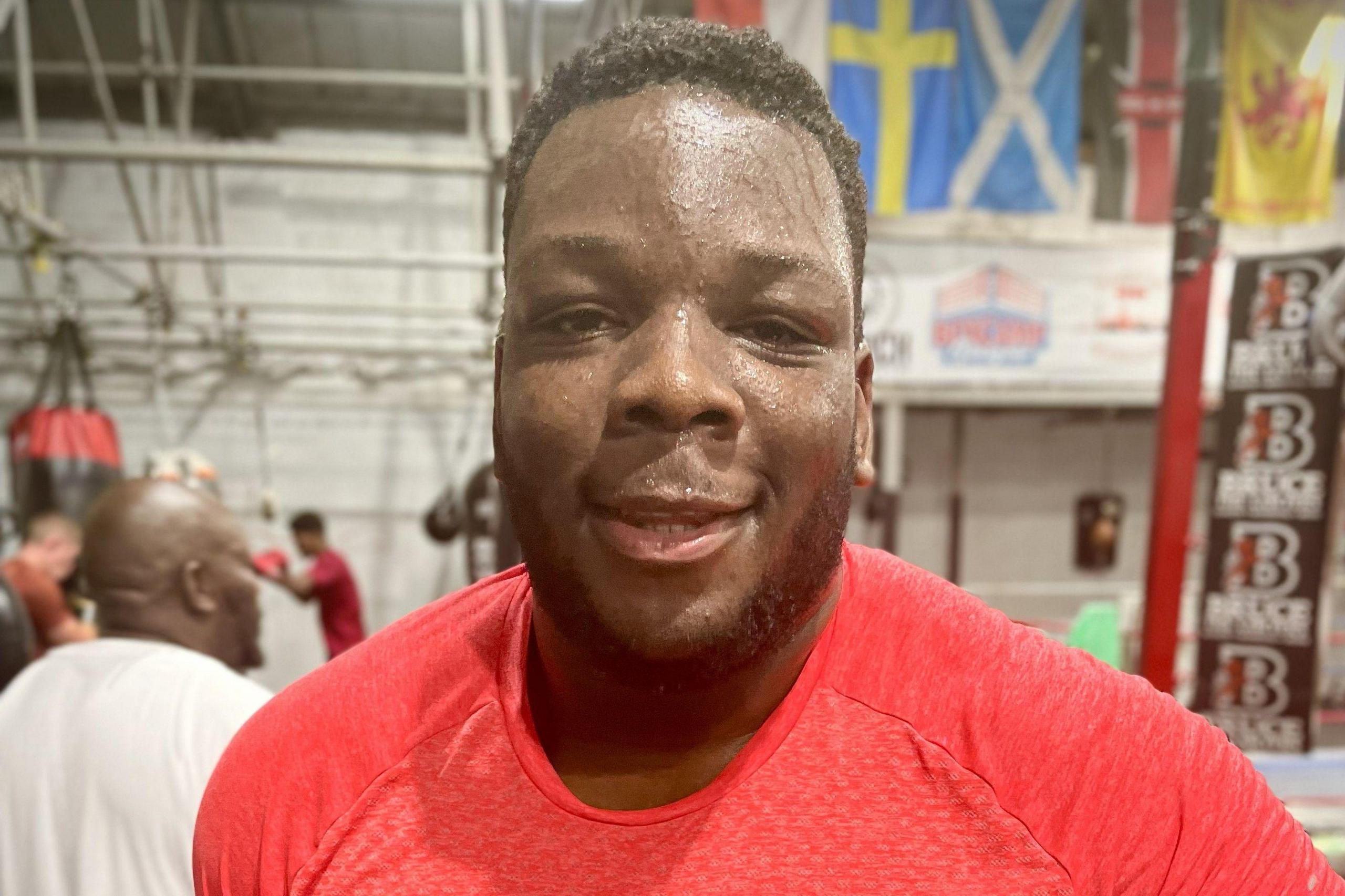 A smiling man with short cropped hair sweats heavily in a gym after boxing training. He is wearing a red t-shirt.