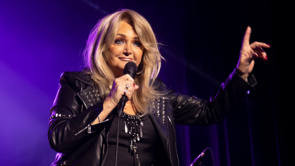 A glamorous woman with blonde hair, wearing all black and holding a microphone, with one hand raised up in the air.