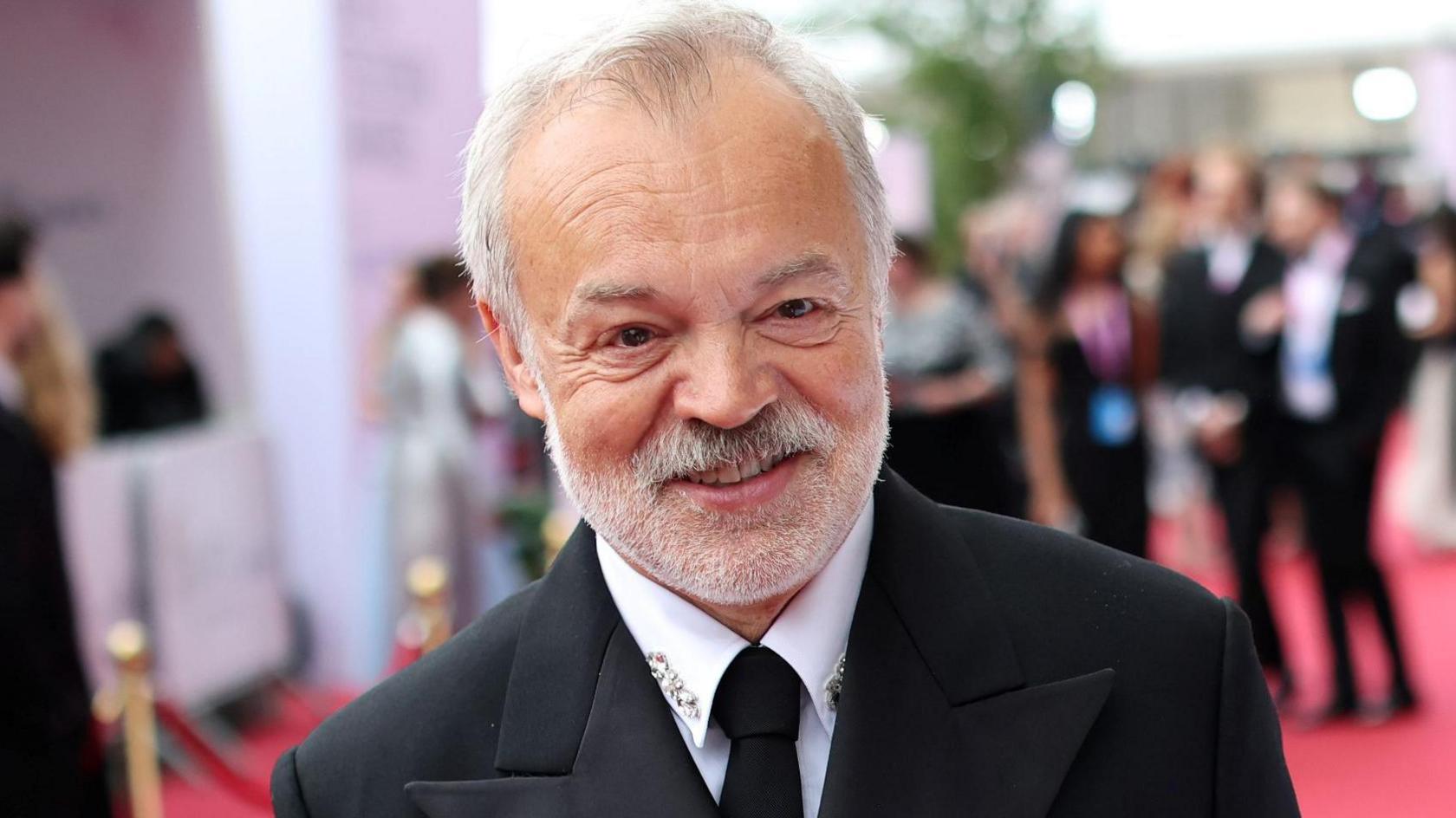 Graham Norton at the Baftas 2026 on the red carpet, wearing a black suit and tie and a white shirt with diamante detail on the collar