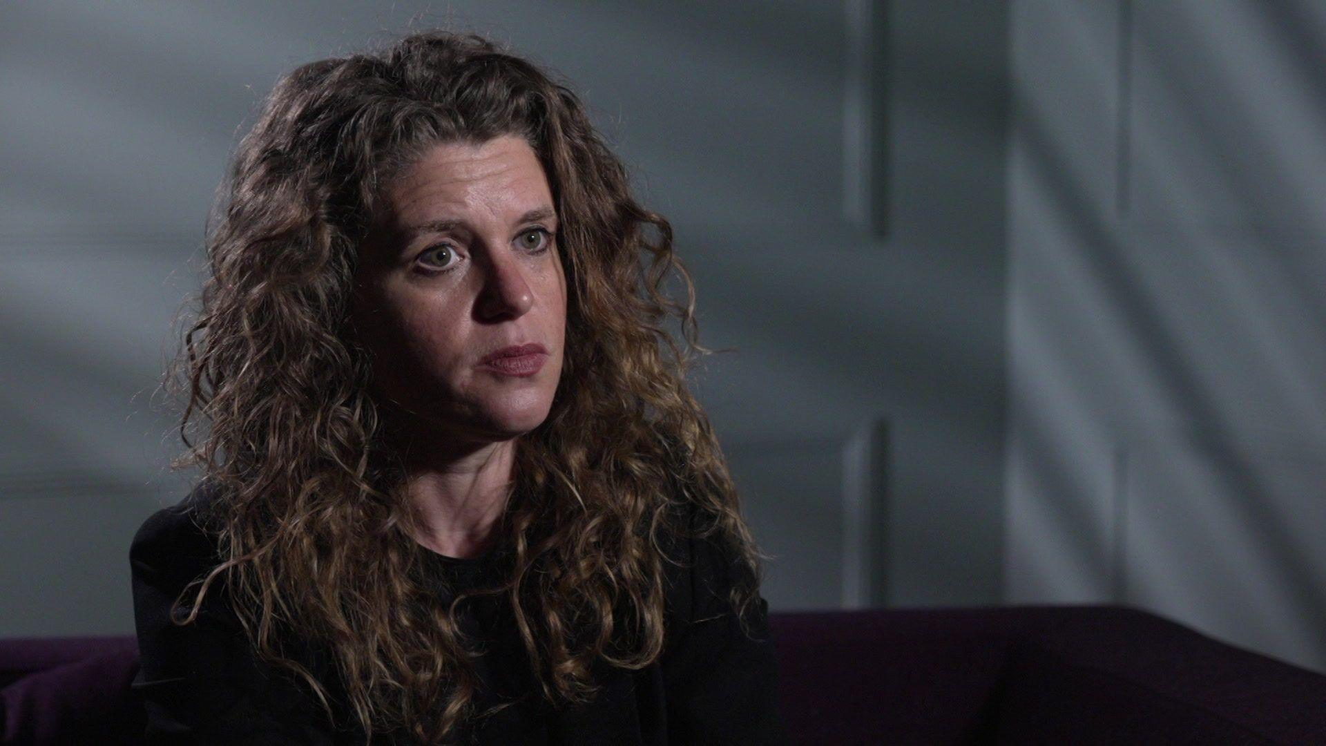 Sophie is sitting inside a room with white walls, she is wearing a black suit and has long, curly hair.