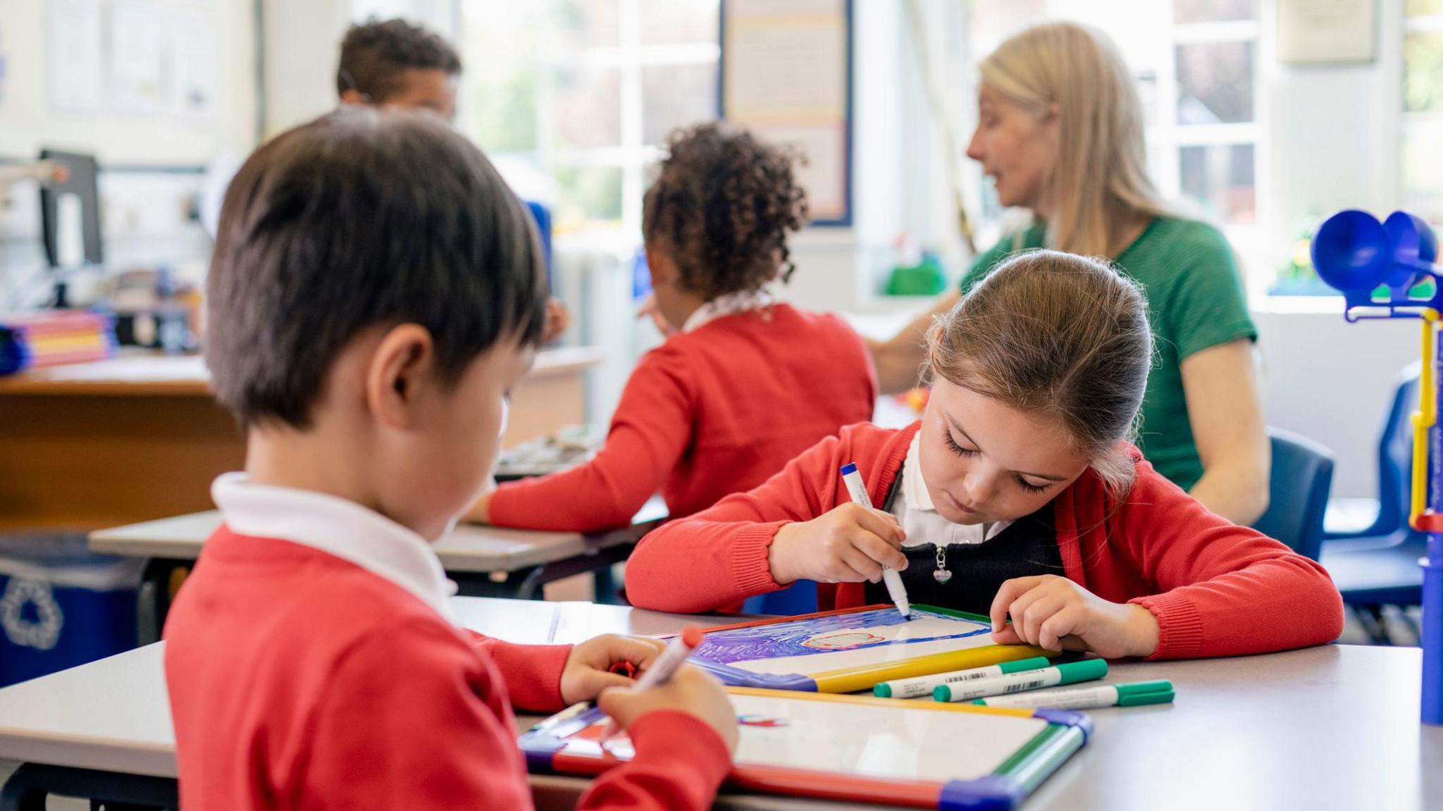 A close up view of a small group of school friends sitting in their classroom. Two pupils at the front of the image are drawing pictures on their whiteboards and a teaching assistant is supervising other students in the background.