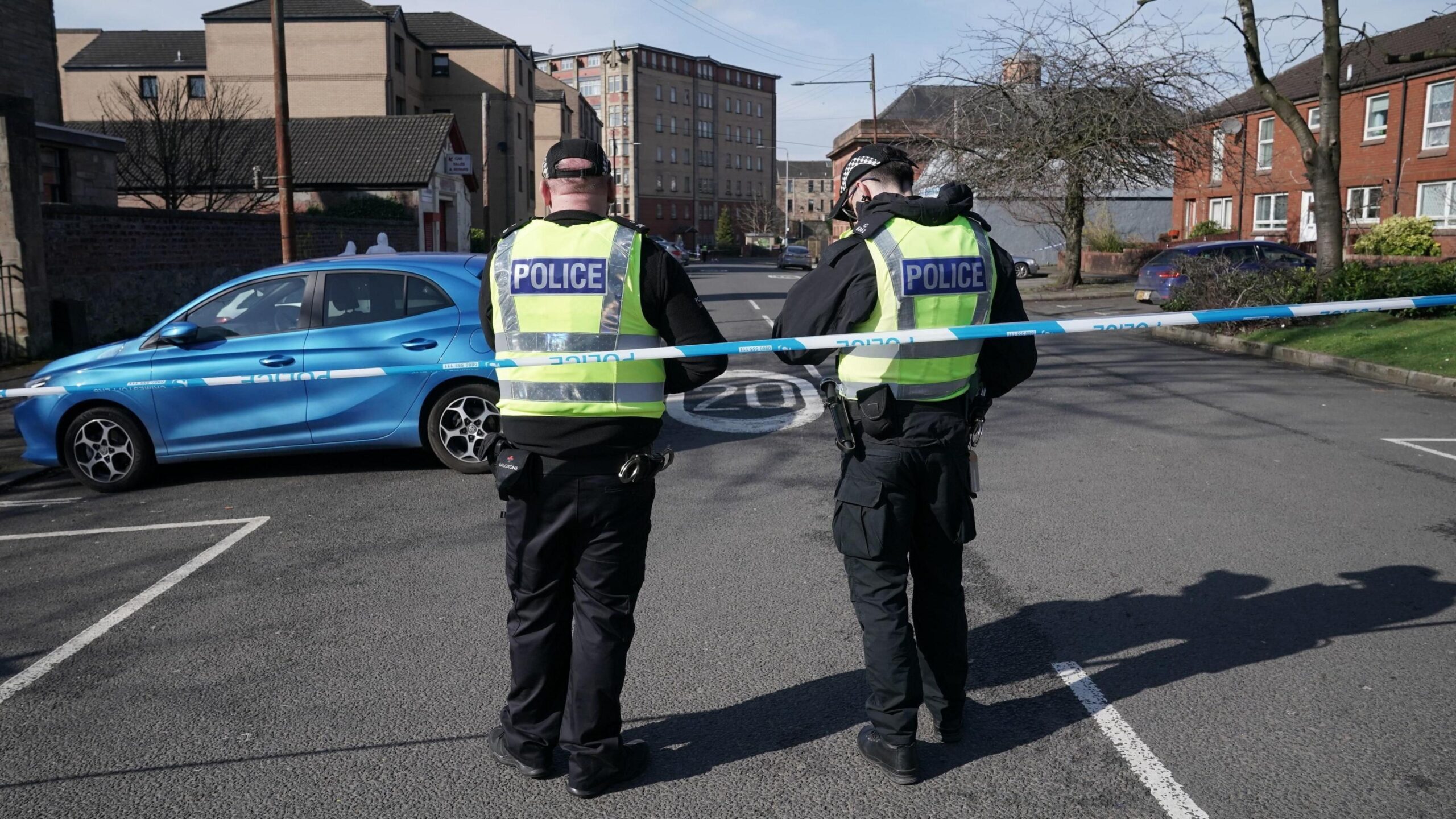 Two police officers stand behind police tape, with their backs to the camera. Flats and houses can be seen in the background, as well as a blue car.
