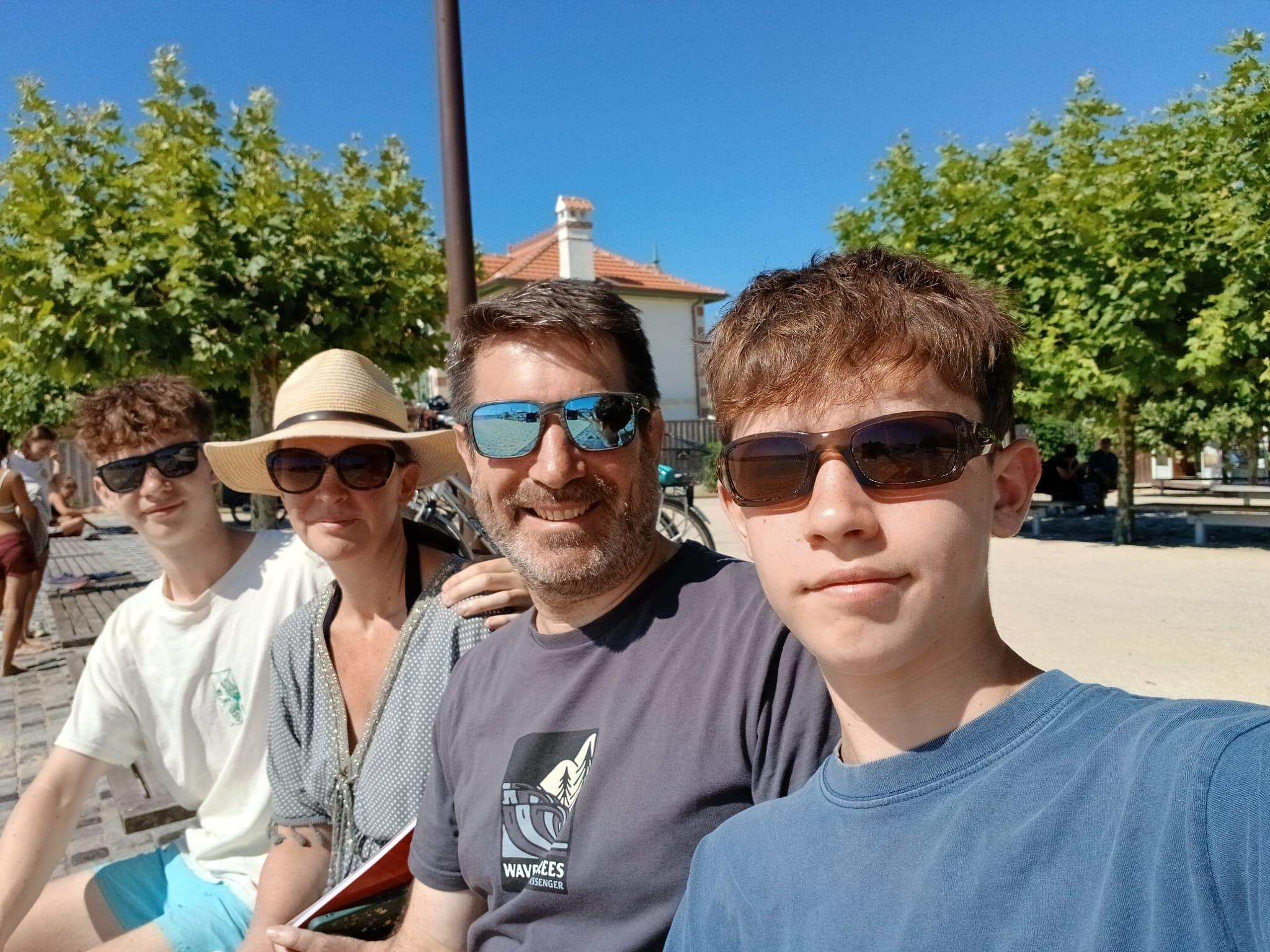 A family picture - two teenage sons are sat either side of their parents with blue sky and trees in the background. All four family members are wearing T-shirts and sunglasses.